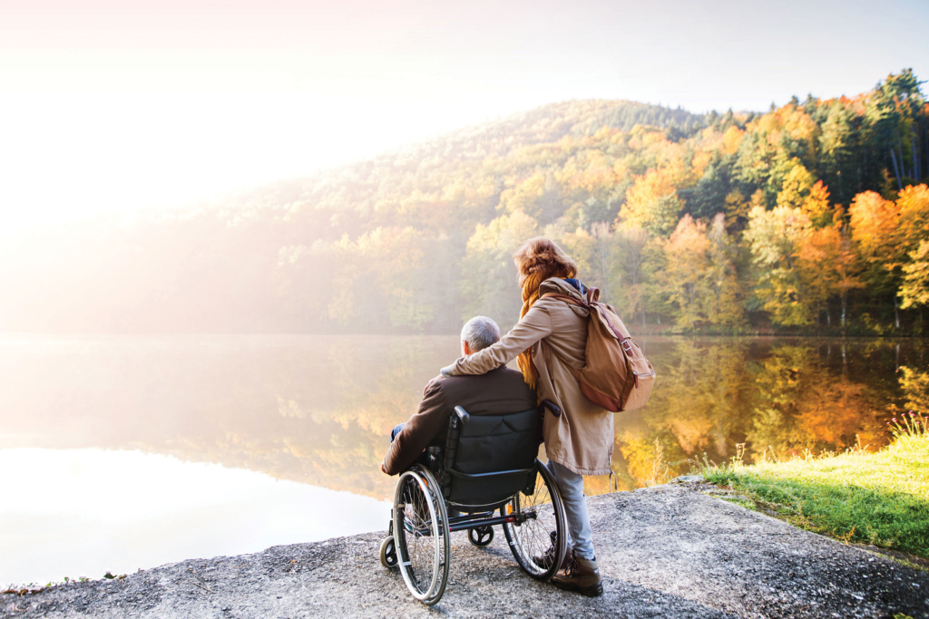 A senior couple stands beside a lake, with one using a wheelchair and the other standing beside them